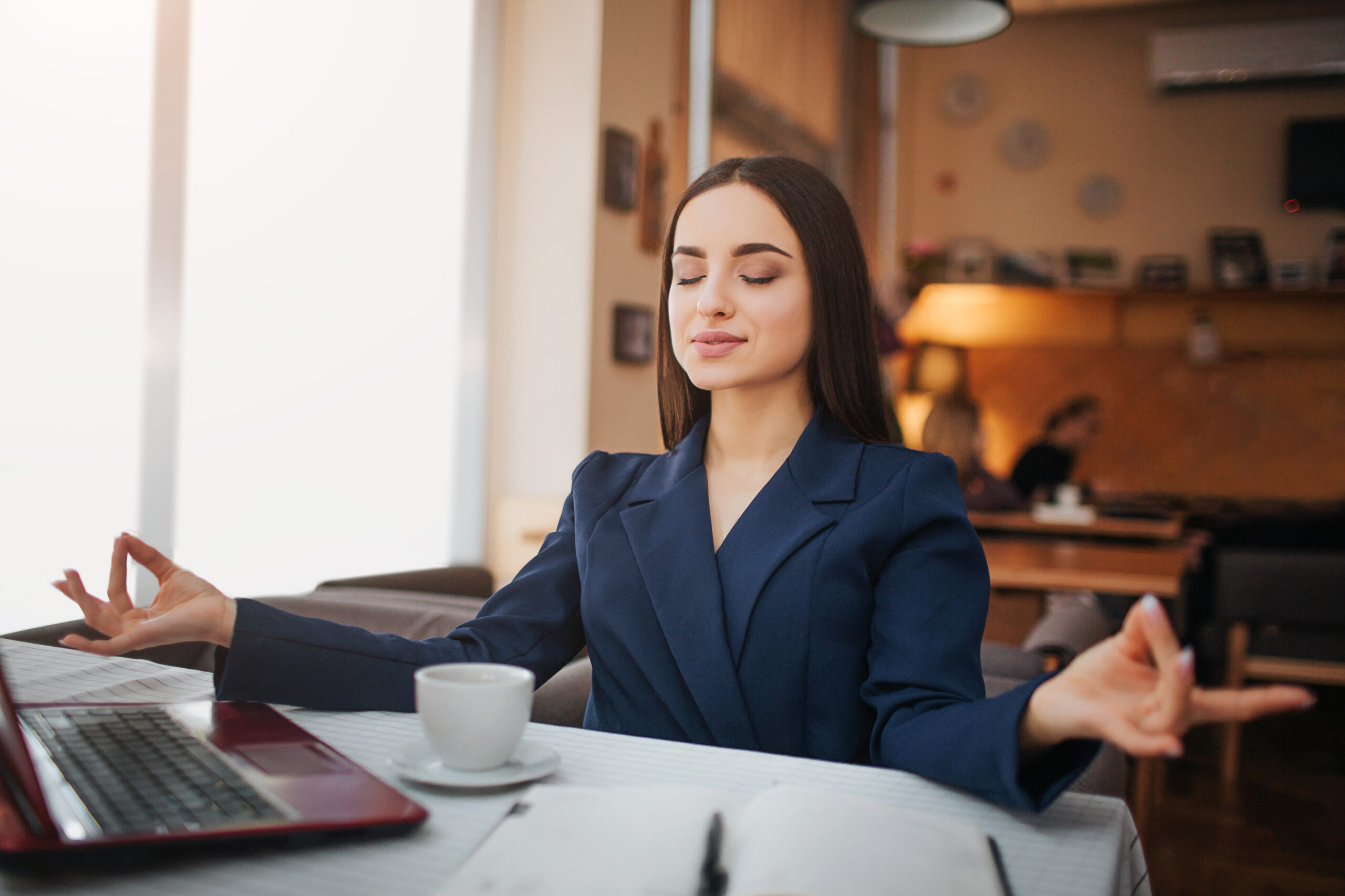 Calm and peaceful young woman sit on table and meditation. She keep eyes closed. Laptop notebook and cup of coffee stand on table